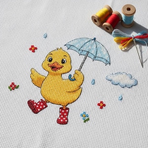 Puede incluir: Bordado de punto de cruz de un patito amarillo con botas de agua rojas con lunares y un paraguas azul. El diseño incluye flores bordadas, gotas de lluvia y una nube. Se ven carretes de hilo y una aguja.