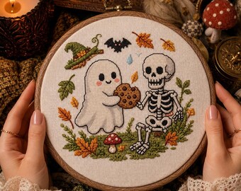 Op de afbeelding: Een ronde houten borduurring met een kruissteekontwerp van een wit spookje en een skelet die samen een koekje met chocoladestukjes eten. Het tafereel bevat een groene heksenhoed, een kleine vleermuis, herfstbladeren en een rode paddenstoel.