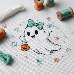 May include: A cross-stitch design of a friendly white ghost with a teal gingham bow and a jack-o'-lantern bucket. The design is surrounded by orange, green, and teal embroidery floss and a needle on a white fabric background. Wooden spools of thread are also visible.