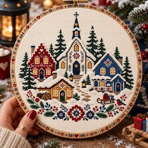 Può includere: Un telaio da ricamo circolare contenente un motivo a punto croce di un villaggio invernale. Il disegno presenta una chiesa bianca centrale, una casa rossa, una blu e una gialla circondate da pini verdi e neve. Un bordo floreale incornicia la parte inferiore.