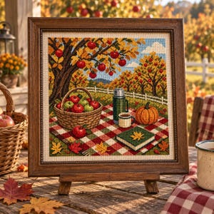 Könnte beinhalten: Ein gerahmtes Kreuzstichbild mit einer herbstlichen Obstgarten-Szene. Ein geflochtener Korb mit roten und grünen Äpfeln steht auf einer rot-weiß karierten Picknickdecke neben einer grünen Thermoskanne, einem Becher, einem Kürbis und einem Buch.