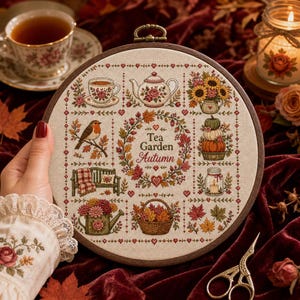 May include: A circular embroidery hoop containing a cross-stitch sampler on beige fabric. The design features the text Tea Garden Autumn surrounded by motifs including a teapot, teacup, robin, pumpkins, sunflowers, a bench, and a basket of leaves.