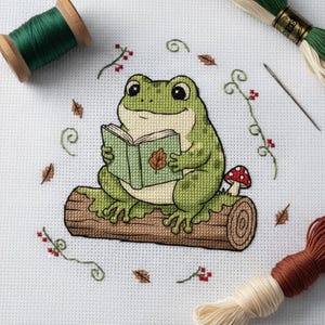 Può includere: Motivo a punto croce con una rana verde che legge un libro su un tronco. La rana è circondata da foglie ricamate, bacche e un fungo. Sono visibili rocchetti di filo da ricamo verde e marrone e un ago.