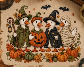 Op de afbeelding: Een kruissteekborduurwerk op beige stof met vier witte eenden in Halloween-kostuums: één met een groene heksenhoed en mantel, één in een pompoen, één met een donkerblauwe sterrenhoed en mantel, en één verkleed als een mummie.