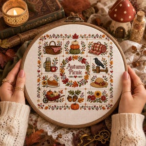 Könnte beinhalten: Ein runder Stickrahmen aus Holz mit einem Kreuzstich-Muster auf weißem Stoff. Das Design zeigt den Schriftzug Autumn Picnic, umgeben von Motiven wie einem Picknickkorb, Kürbissen, einer Thermoskanne, einer Krähe, einem Stück Kuchen und Blumen.
