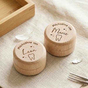 Könnte beinhalten: Zwei kleine, runde Holzkästchen mit Deckeln. Die Deckel sind mit den Worten "Milchzähne von Marie" und "Milchzähne von Leon" sowie einer Zahnzeichnung graviert. Die Kästchen sind hellbraun und liegen auf einer neutralfarbenen Oberfläche.
