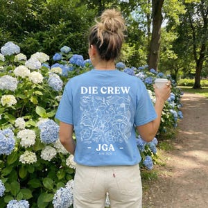 May include: A light blue t-shirt with the text "DIE CREW" and a floral design on the back. Below the text is "- JGA -" and "JUNI 2026". The person is standing in front of a bush of blue and white hydrangeas.