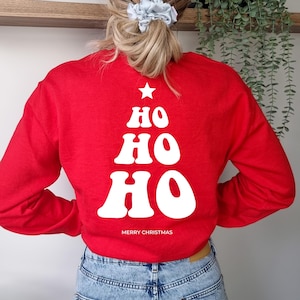 Könnte beinhalten: Ein leuchtend rotes Langarm-Sweatshirt mit einer weißen Grafik auf der Rückseite. Das Design ist wie ein Weihnachtsbaum geformt, mit dem Text HO HO HO in einer Retro-Schrift, einem kleinen Stern an der Spitze und MERRY CHRISTMAS darunter.
