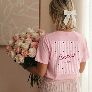 Ein rosa T-Shirt mit dem Wort "Crew" und dem Datum "Est. 2026" auf der Rückseite, mit einem gestreiften Muster und Herzen. Die Person hält einen großen Strauß rosa und weißer Rosen. Die Person trägt eine gestreifte Hose.