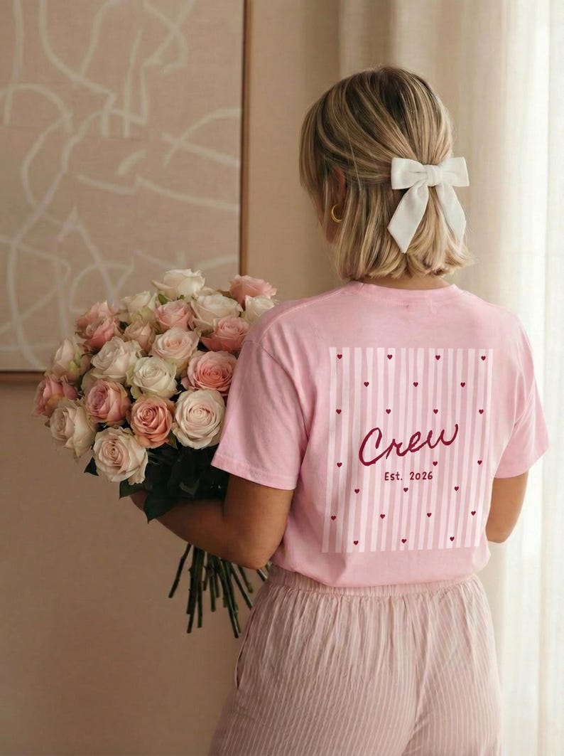Ein rosa T-Shirt mit dem Wort "Crew" und dem Datum "Est. 2026" auf der Rückseite, mit einem gestreiften Muster und Herzen. Die Person hält einen großen Strauß rosa und weißer Rosen. Die Person trägt eine gestreifte Hose.