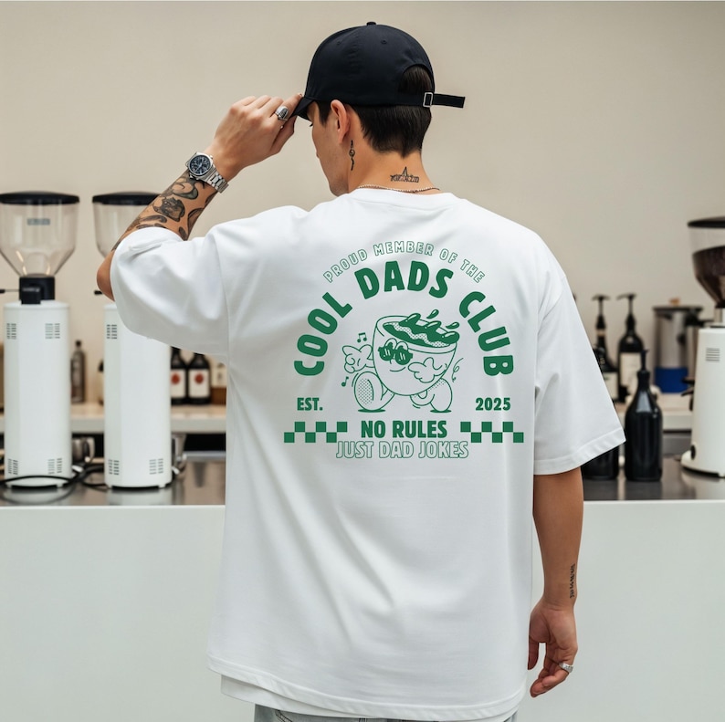 May include: White t-shirt with a green graphic on the back that reads "COOL DADS CLUB" with a cartoon image of a bowl. The text "EST. 2025" and "NO RULES JUST DAD JOKES" are also printed on the shirt.