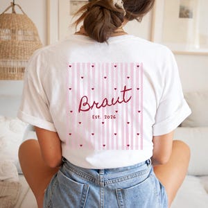 May include: White t-shirt with a pink and white striped design on the back, adorned with small red hearts. The word "Braut" is written in a cursive font, with "Est. 2026" below it. The shirt is a casual style.