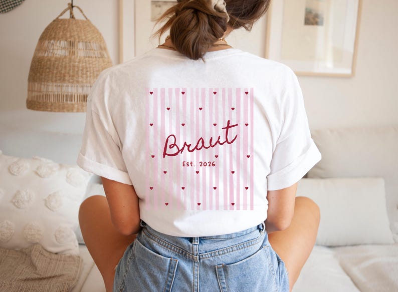 May include: White t-shirt with a pink and white striped design on the back, adorned with small red hearts. The word "Braut" is written in a cursive font, with "Est. 2026" below it. The shirt is a casual style.