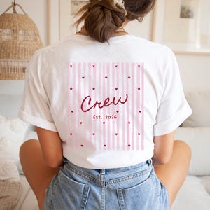May include: White t-shirt with a pink and white striped design on the back, featuring the word "Crew" in a cursive font, with small heart accents and the year "Est. 2026". The shirt is a casual, everyday wear item.