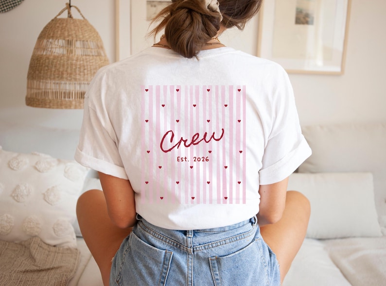 May include: White t-shirt with a pink and white striped design on the back, featuring the word "Crew" in a cursive font, with small heart accents and the year "Est. 2026". The shirt is a casual, everyday wear item.
