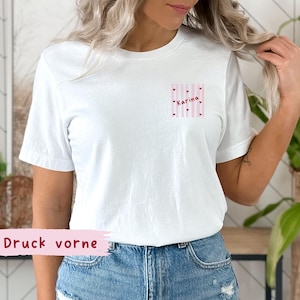 May include: White t-shirt with a small pocket design featuring pink and white stripes, small hearts, and the name "Karina" in pink. The text "Druck vorne" is printed on a pink banner at the bottom.