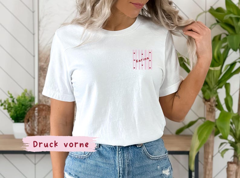 May include: White t-shirt with a small pocket design featuring pink and white stripes, small hearts, and the name "Karina" in pink. The text "Druck vorne" is printed on a pink banner at the bottom.