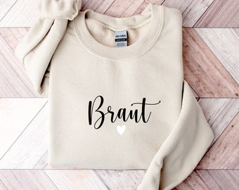 May include: A beige sweatshirt with the word "Braut" in black cursive lettering and a white heart below it.