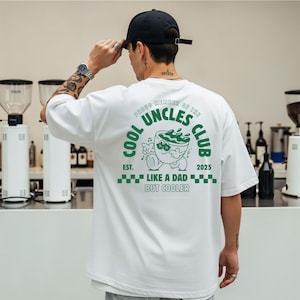 Könnte beinhalten: Weißes T-Shirt mit einem grünen Aufdruck auf der Rückseite mit der Aufschrift "Cool Uncles Club" und einem Cartoon-Design. Der Text "Like a Dad But Cooler" ist ebenfalls enthalten. Das T-Shirt wird von einer Person mit einer schwarzen Kappe getragen.
