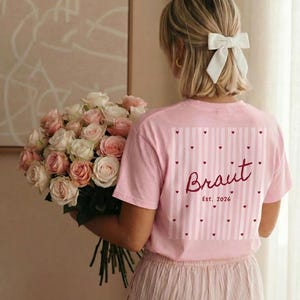 May include: Pink t-shirt with the word "Braut" and "Est. 2026" printed on the back, along with a striped and heart design. The person is holding a large bouquet of pink and white roses. The person is wearing a white bow.