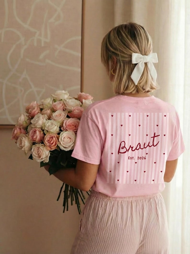 May include: Pink t-shirt with the word "Braut" and "Est. 2026" printed on the back, along with a striped and heart design. The person is holding a large bouquet of pink and white roses. The person is wearing a white bow.