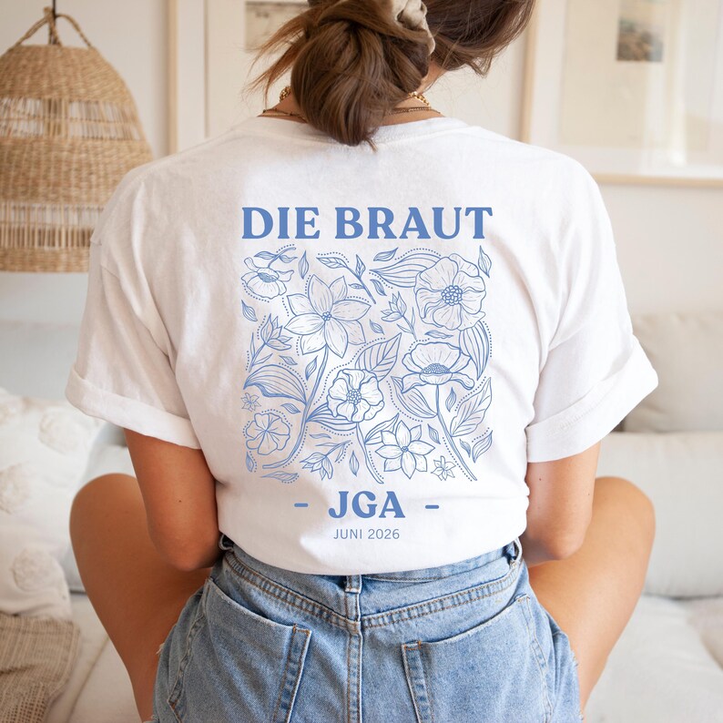 May include: White t-shirt with a blue floral design and the text "DIE BRAUT - JGA - JUNI 2026". The design features various flowers and leaves. The shirt is made of a soft material.
