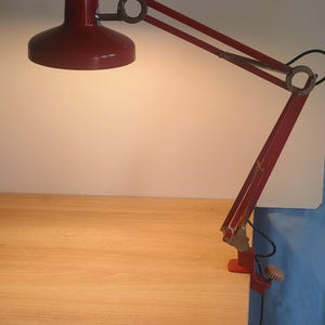 May include: A red desk lamp with an adjustable arm and a clamp base. The lamp features a rounded shade and a flexible arm, enabling directional lighting. The lamp is made of metal and is designed to be attached to a desk or table.