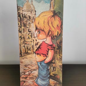 May include: Vintage rectangular art print featuring a cartoon child with blonde hair, wearing a red shirt and blue jeans, smoking a cigarette. The background depicts a European cityscape with a church. The artwork is on a dark brown surface.