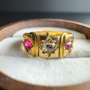 Puede incluir: Un anillo de oro con tres piedras engastadas en la banda. La piedra central es un diamante transparente y las dos piedras laterales son rubíes rojos.