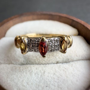 May include: A gold ring featuring three marquise-shaped gemstones. The central stone is red, while the two outer stones are yellow. The band is set with small, clear stones.