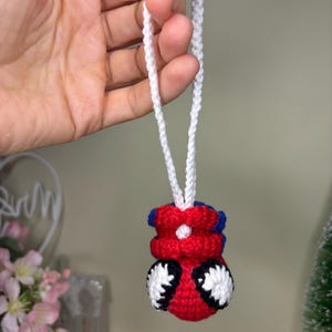 May include: A handmade, crocheted Spider-Man ornament. The ornament is mainly red with black and white accents, and blue details. It hangs from a white crocheted cord. The ornament is approximately 5 cm tall.