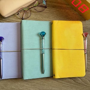 May include: Three rectangular notebooks in lavender, mint green, and yellow with textured covers. Each notebook is held closed by a thin elastic band and features a matching ballpoint pen with a large, faceted, diamond-shaped topper.
