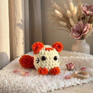 Puede incluir: Un juguete de tigre hecho a mano, tejido a ganchillo en crema y naranja. El peluche tiene grandes ojos negros, orejas, patas y cola naranjas. El tigre está sentado sobre una superficie blanca texturizada. También se ven un anillo de madera y flores secas.