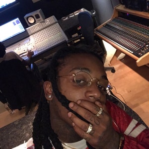 May include: A man with long black dreadlocks is wearing a red and white striped shirt, a gold chain, and a gold watch. He is wearing gold-rimmed glasses and has his hand over his mouth. He is in a recording studio.