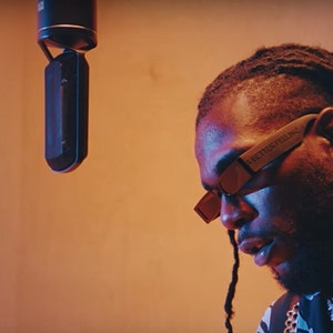 Peut inclure: Une personne portant des lunettes de soleil avec le texte "A BETTER FEELING" sur le côté, avec un microphone en arrière-plan. La personne porte une chemise à motifs et un collier.