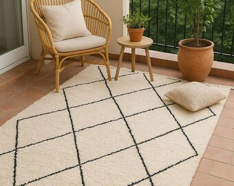 May include: Cream-colored rug with a black diamond pattern and fringe trim. The rug is placed on a brick patio, alongside a wicker chair with a cream cushion, a small wooden table, and potted plants. A matching cream pillow rests on the rug.