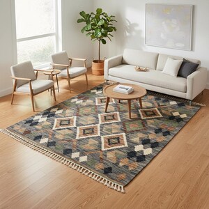 May include: A rectangular area rug with a geometric diamond pattern in shades of gray, brown, green, and cream. The rug has a fringe on the short ends and is placed in a living room setting with furniture.