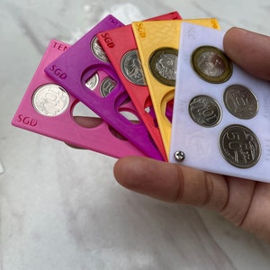 May include: A set of five coin holders, each with a different color: purple, pink, red, yellow, and white. Each holder has a circular cutout for a coin and the text "SGD" printed on it.
