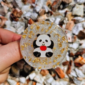 May include: A white and black panda bear with a red apple in its mouth is featured on a round, clear resin coaster with gold glitter and flecks.