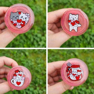 May include: Four pink resin cat-themed round ornaments. Each ornament features a different design with cats and hearts. The first ornament features two cats with hearts around them. The second ornament features a cat holding a bunch of hearts. The third ornament features two cats in a teacup with hearts around them. The fourth ornament features a cat in a jar with hearts around it.