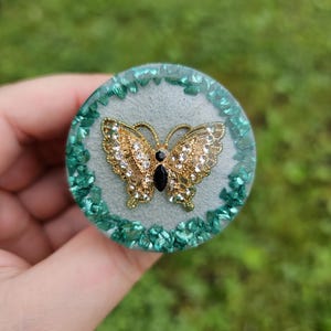 May include: A round, light blue resin paperweight with a gold butterfly design. The butterfly has clear rhinestones and a black centre. The paperweight is surrounded by a border of green glitter.