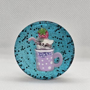 May include: A white cat with a pink nose and whiskers is sitting in a purple teacup with white polka dots. The cat is wearing a red strawberry on its head. The teacup is on a blue background with black glitter.