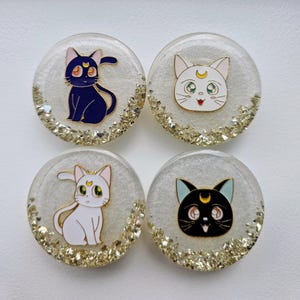 May include: Four round, white, resin phone grips with gold glitter. Each grip features a different cartoon cat with a crescent moon and star design. The cats are black, white, and black and white.