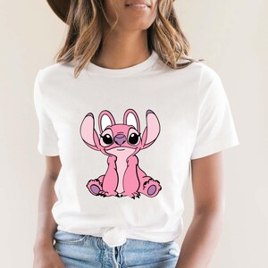 May include: White t-shirt featuring a pink cartoon character with large ears and a friendly expression. The character is sitting, with its arms and legs visible. The design is centered on the shirt.
