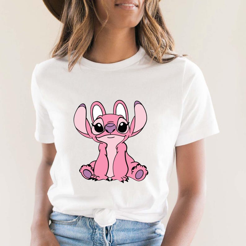 May include: White t-shirt featuring a pink cartoon character with large ears and a friendly expression. The character is sitting, with its arms and legs visible. The design is centered on the shirt.