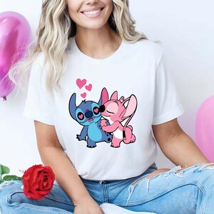 May include: White t-shirt featuring a cartoon illustration of two characters embracing, with hearts floating above them. One character is blue, the other is pink. The design is centered on the shirt.