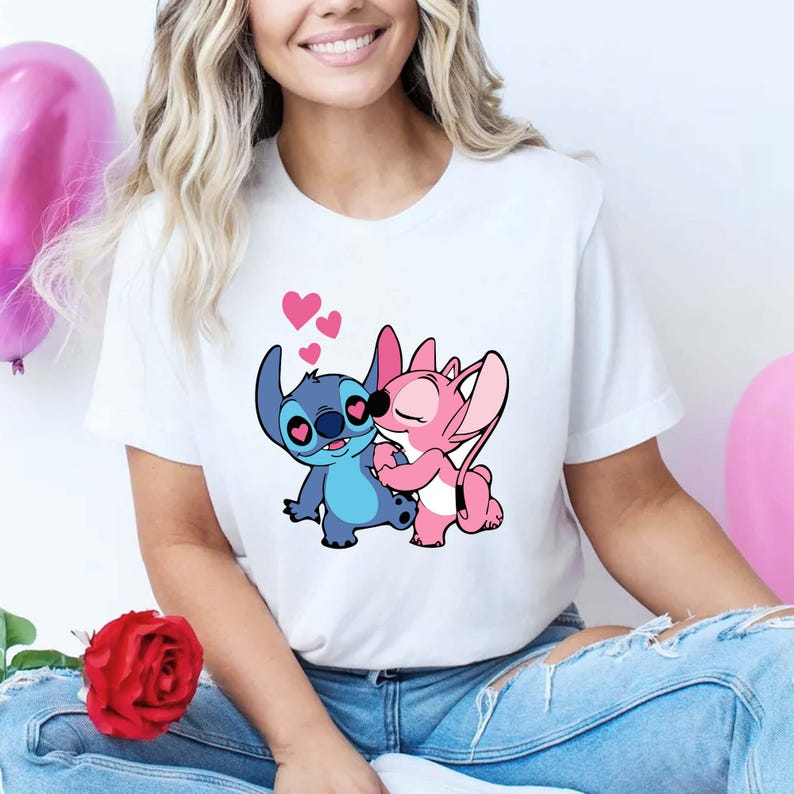 May include: White t-shirt featuring a cartoon illustration of two characters embracing, with hearts floating above them. One character is blue, the other is pink. The design is centered on the shirt.