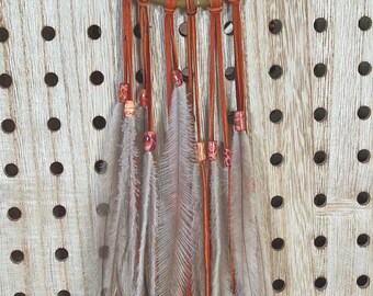 May include: A handcrafted dreamcatcher with a circular woven frame, adorned with feathers and beads. The dreamcatcher features a tan-colored woven center, orange leather strips, and gray feathers. It hangs against a light wood background.
