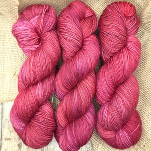 Puede incluir: Tres madejas de hilo teñido a mano en un vibrante color rosa magenta. Cada madeja está retorcida en una bobina larga y gruesa, mostrando un tono rico y saturado con variaciones sutiles y matices púrpuras oscuros en las fibras retorcidas.