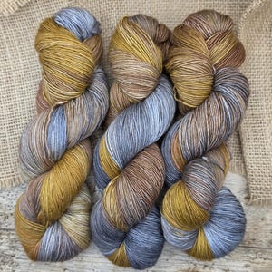 Op de afbeelding: Drie strengen handgeverfd garen in een mix van kleuren. Het garen heeft tinten goud, bruin en blauw, waardoor een gemêleerd effect ontstaat. Het garen is gerangschikt op een houten oppervlak met een jute achtergrond.